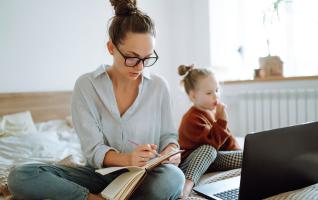Frau macht sich zu Hause Notizen in einem Heft, neben ihr ein Kind und ein aufgeklappter Laptop.