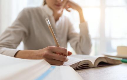 Person schreibt an einem Tisch in ein Heft, daneben liegt ein aufgeschlagenes Buch.