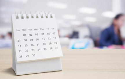 Weißer Tischkalender mit Monatsübersicht auf einem Schreibtisch vor einem unscharfen Büro.