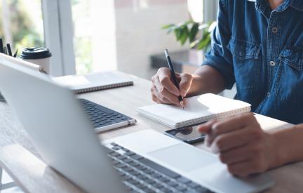 Eine Person macht sich neben dem Laptop Notizen und prüft vor der Anmeldung die Angaben eines Online-Kurses zu Umfang und Prüfungsform