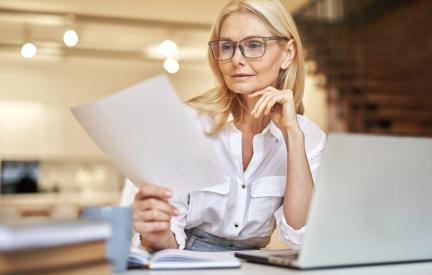 Frau am Schreibtisch prüft ein Dokument neben ihrem aufgeklappten Laptop